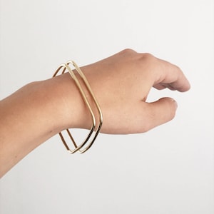 May include: Two gold square-shaped bangles stacked on a person's wrist.