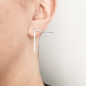 May include: A silver ear cuff with a geometric design. The cuff is shaped like a right angle, with a long vertical bar and a shorter horizontal bar.