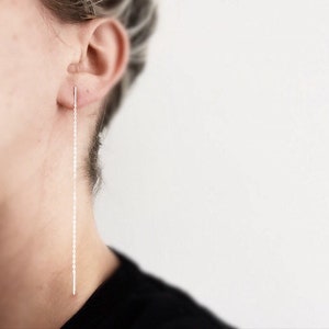 Puede incluir: Un pendiente largo y delgado de cadena plateada cuelga del lóbulo de la oreja de una persona.