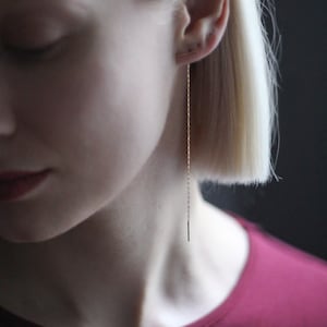 May include: A close-up of a person wearing a long, thin gold chain earring with a small bar at the end. The earring is hanging down the side of their neck.