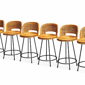 May include: A set of six bar stools featuring round woven rattan seats and curved rattan backrests. Each stool is supported by four slender black metal legs connected by a circular footrest ring, providing a stable base for the natural wood-toned seats.