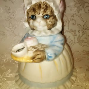 May include: A ceramic figurine of a cat wearing a pink bonnet and blue dress, holding a tray with a cup and a pink cupcake.
