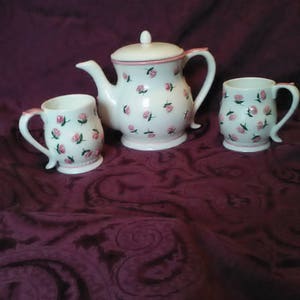 May include: A white teapot and two mugs with pink and green floral designs. The teapot has a lid and a handle. The mugs have handles. The set is on a dark purple background.