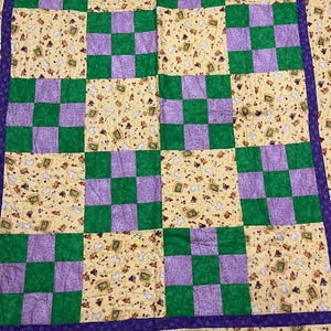 May include: A patchwork quilt with a checkerboard pattern. The quilt features alternating squares of green and purple, set against a yellow background with a repeating pattern of small teddy bears and picture frames. A purple border frames the quilt.