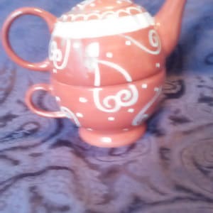 May include: A coral-colored ceramic tea set with a teapot and teacup. The teapot has a lid and spout, and the teacup sits beneath it. Both pieces are decorated with white swirls and dots.