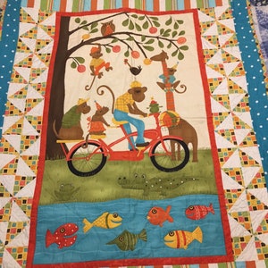 May include: A colorful quilt with a whimsical design featuring monkeys riding a bicycle, a giraffe, and fish swimming in a blue stream. The quilt has a white background with a blue, orange, and green border.