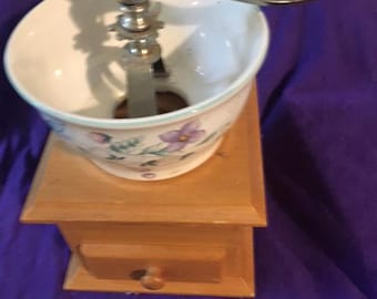 May include: A vintage wooden coffee grinder with a porcelain top featuring a floral design. The grinder has a hand crank and a drawer for collecting coffee grounds.