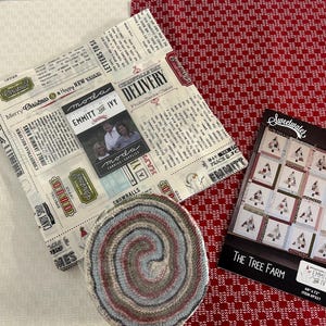 May include: A fabric bundle with a newspaper print, a rolled fabric, and a pattern booklet. The fabric bundle features text and holiday-themed graphics. The rolled fabric has a spiral pattern in shades of blue, gray, and red. The booklet is titled "The Tree Farm."