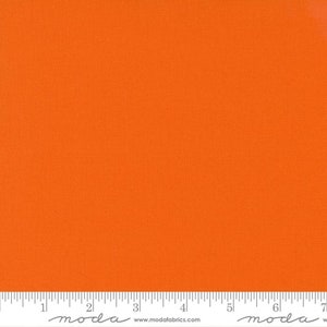 May include: Bright orange fabric with a textured surface. A ruler is at the bottom of the image, with the word "moda" and the website address "www.modafabrics.com" printed on it. The fabric is suitable for quilting and sewing projects.