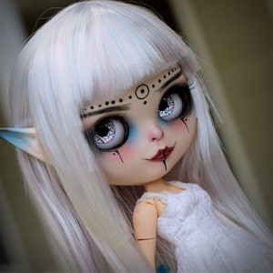 Puede incluir: Una mu&ntilde;eca blanca con el pelo largo, liso y blanco. La mu&ntilde;eca tiene maquillaje de ojos azul y negro, una boca roja y orejas de elfo. La mu&ntilde;eca lleva un vestido blanco.