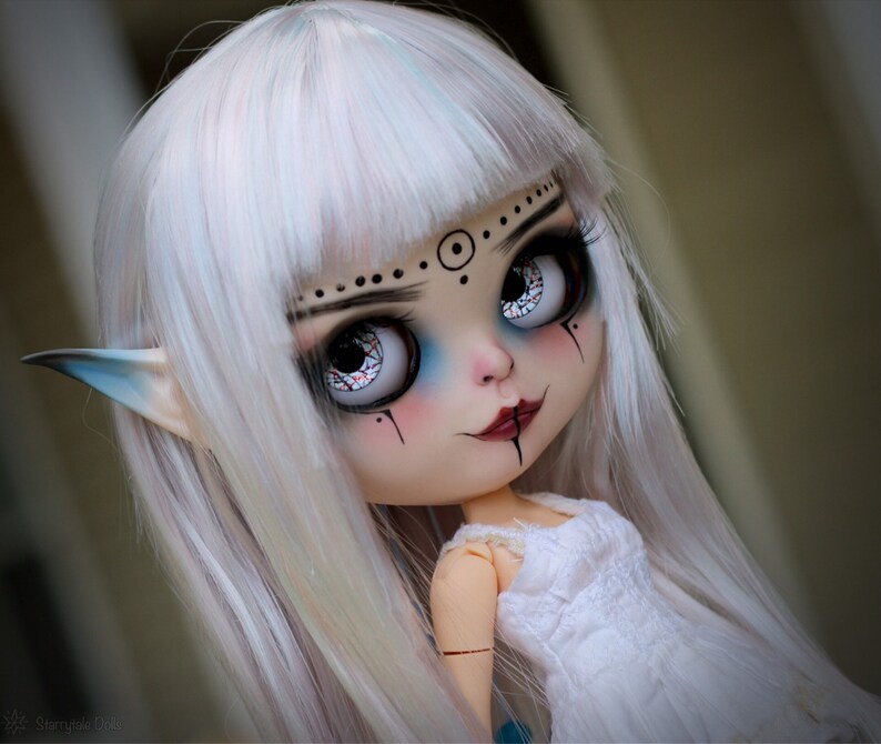 Puede incluir: Una mu&ntilde;eca blanca con el pelo largo, liso y blanco. La mu&ntilde;eca tiene maquillaje de ojos azul y negro, una boca roja y orejas de elfo. La mu&ntilde;eca lleva un vestido blanco.