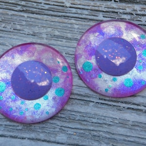 May include: Two round, purple and pink earrings with a central, glittery, light purple circle. The earrings have a shimmering, iridescent effect with teal-colored dots. The earrings are set against a weathered, wooden surface.