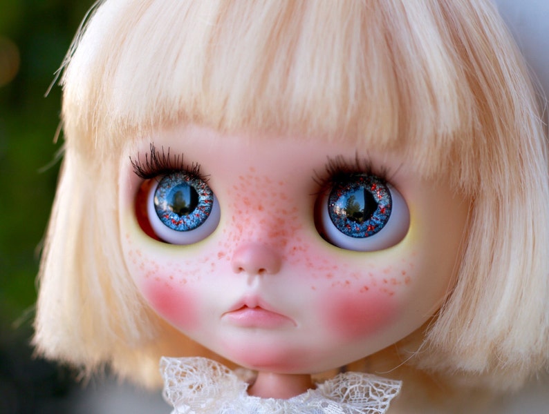 Puede incluir: Primer plano de la cara de una mu&ntilde;eca con ojos azules con purpurina roja, cabello rubio claro y mejillas sonrosadas.