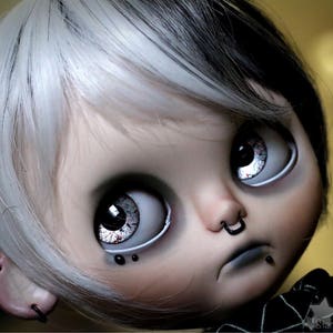 Puede incluir: Una mu&ntilde;eca con cabello blanco y negro, ojos grandes azules y un aro de nariz negro. La mu&ntilde;eca lleva una camisa a rayas blancas y negras y tiene un aro de labio negro.