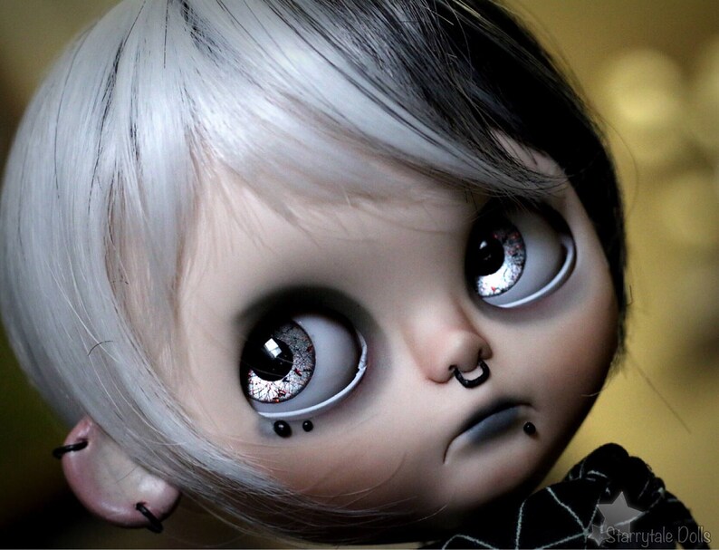 Puede incluir: Una mu&ntilde;eca con cabello blanco y negro, ojos grandes azules y un aro de nariz negro. La mu&ntilde;eca lleva una camisa a rayas blancas y negras y tiene un aro de labio negro.