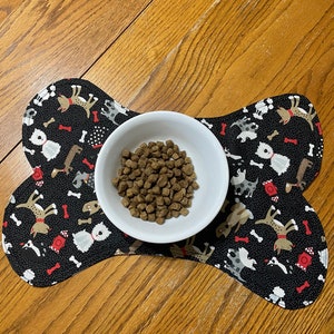 May include: A black bone-shaped dog bowl mat with a white bowl filled with brown dog food. The mat is covered in a pattern of dogs and bones.