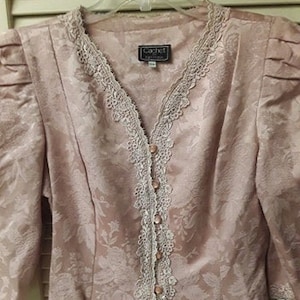 May include: A blush pink button-up blouse with a floral pattern and lace trim. The blouse has a V-neckline and long sleeves. The label reads "Cachet Boutique".
