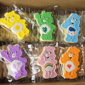 May include: A box of frosted sugar cookies shaped like Care Bears. The cookies are individually wrapped and feature various colors, including yellow, green, blue, purple, pink, and orange. Each bear has a unique belly badge, such as a sun, a four-leaf clover, or a rainbow.