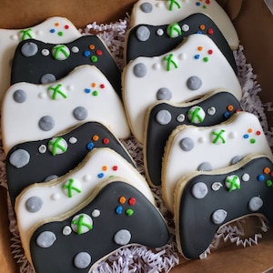 May include: A box of decorated sugar cookies shaped like video game controllers. The cookies are white and black with green, red, and blue details.