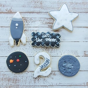 May include: A set of six decorated sugar cookies with a space theme. The cookies are decorated with black and white icing and feature a rocket, a star, a moon, a planet, a number two, and the words "Two the Moon".