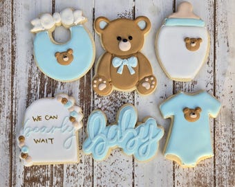 May include: A selection of baby-themed biscuits, including a teddy bear, bib, baby bottle, onesie, and cookies spelling "baby" and "We can bearly wait." The biscuits are decorated with blue and brown icing.