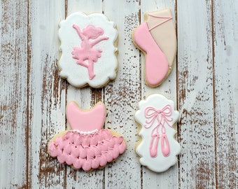 Op de afbeelding: Vier suikerkoekjes met balletthema op een wit houten oppervlak. De koekjes bevatten een ballerina, een balletschoen, een tutu en spitsschoenen. Elk koekje is versierd met roze en witte glazuur.