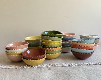 May include: A collection of small ceramic bowls with scalloped edges, arranged in multiple stacks. The bowls feature a variety of solid colors including yellow, pink, green, blue, and brown, with a smooth, glazed finish on the interior surfaces.