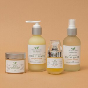 May include: Four bottles of organic skincare products with orange blossom scent. The products include a facial cleanser, facial toner, facial serum, and cleansing balm. All products are labeled "Organic Pure Creations" and are handmade in Georgia.
