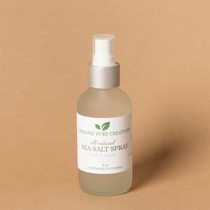 May include: A frosted glass bottle with a silver cap and white spray nozzle. The label reads "Organic Pure Creations all natural Sea Salt Spray with lavender, 4 oz, Handmade in Georgia."