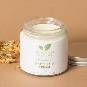 May include: A glass jar of all-natural diaper rash cream, labeled "Organic Pure Creations", with a green leaf logo. The jar is 4 oz and handmade in Georgia.