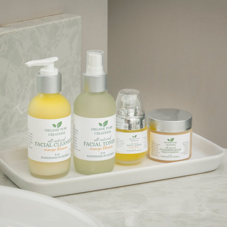 May include: Four bottles of organic facial care products in a white tray. The bottles are labeled "Organic Pure Creations" and contain facial cleanser, facial toner, facial serum, and cleansing balm. The products are all natural and made with orange blossom.