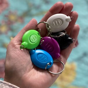 May include: A collection of small, colorful LED keychain flashlights in green, blue, purple, black, and white. Each flashlight has a small, clear light at the end and a silver keyring. The flashlights are held in a hand.