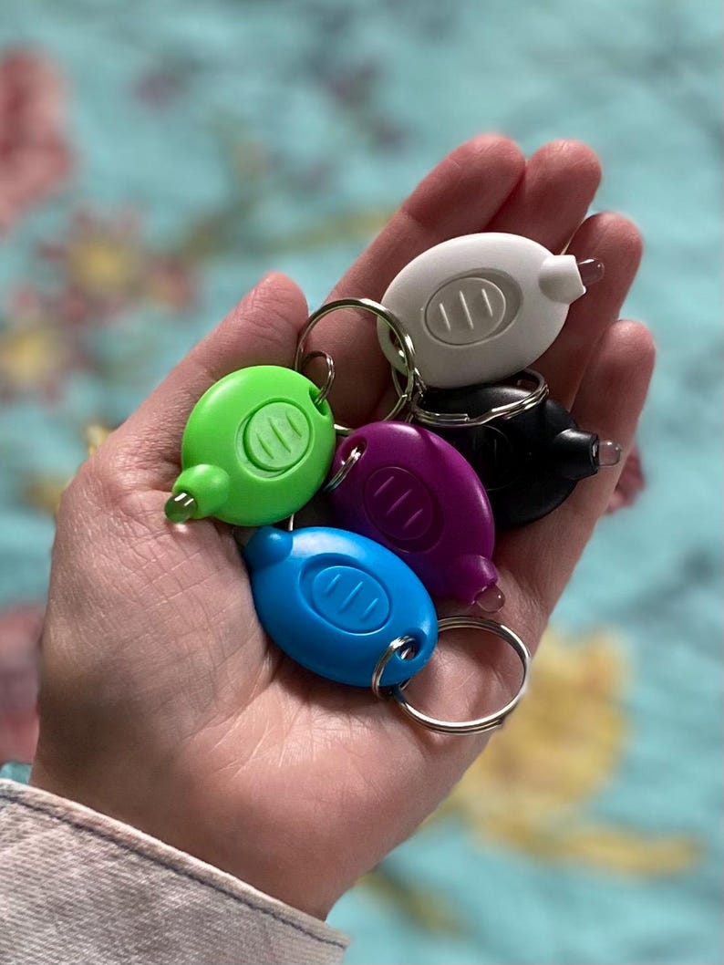 May include: A collection of small, colorful LED keychain flashlights in green, blue, purple, black, and white. Each flashlight has a small, clear light at the end and a silver keyring. The flashlights are held in a hand.