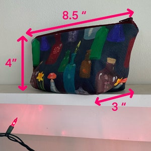 May include: A zippered pouch with a dark blue background and a colorful bottle design. The pouch measures 8.5 inches long, 4 inches high, and 3 inches wide. The zipper is a dark burgundy color.