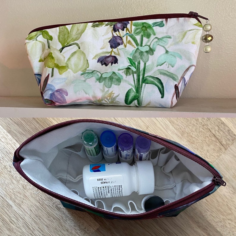 May include: A floral print cosmetic bag with a burgundy zipper and a beaded zipper pull. The bag is open, revealing small bottles and a white pill bottle. The interior is white with pockets.