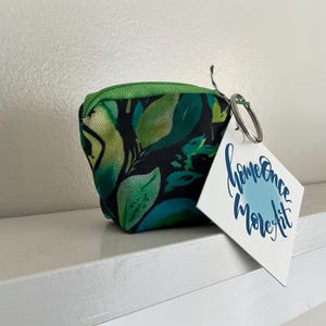 May include: A small fabric coin pouch featuring a dark green and lime leaf pattern with a bright green zipper. A silver metal keyring is attached to the side, holding a white rectangular tag with the blue text Home Once More Art printed on it.