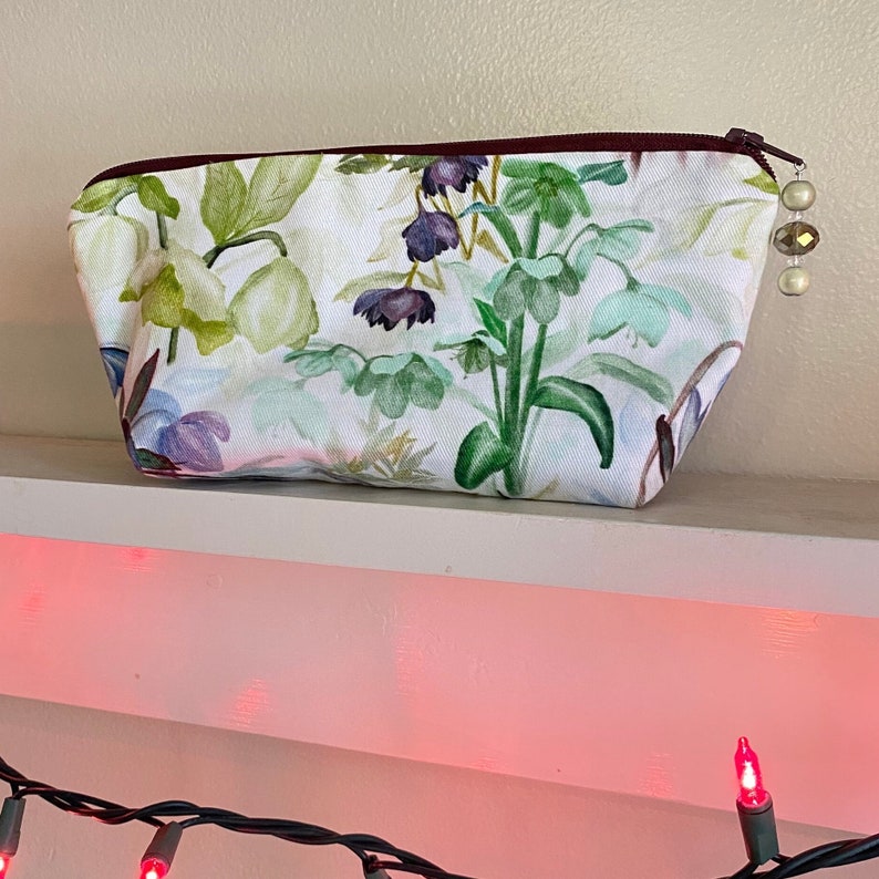 May include: A white cosmetic bag with a floral print featuring green, purple, and blue flowers. The bag has a burgundy zipper and a decorative beaded pull. The bag is sitting on a white shelf.