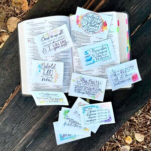 以下が含まれることがあります： 水彩画のデザインが施された、カラフルで手描きの聖書の詩のカードのコレクションです。カードには、「疲れて重荷を負っている人は、みんなわたしのところに来なさい。わたしがやすませてあげます。」や「見よ、わたしはすべてを新しくする。」などのインスピレーションを与える引用が書かれています。
