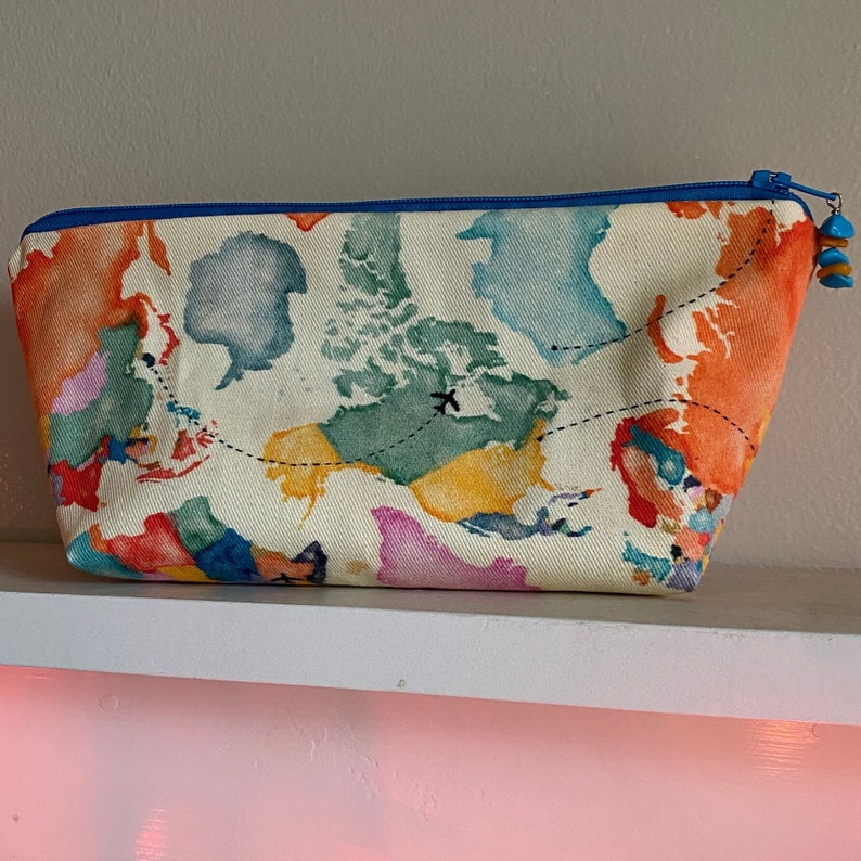 May include: A rectangular cosmetic bag with a world map design. The bag features a blue zipper and a blue and turquoise charm. The map is in watercolor style with orange, green, blue, and pink continents on a cream background.