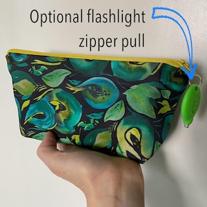 May include: A small, rectangular pouch with a floral pattern in shades of green and teal on a black background. The zipper is yellow, and the optional flashlight zipper pull is green. The text "Optional flashlight zipper pull" is above the pouch.