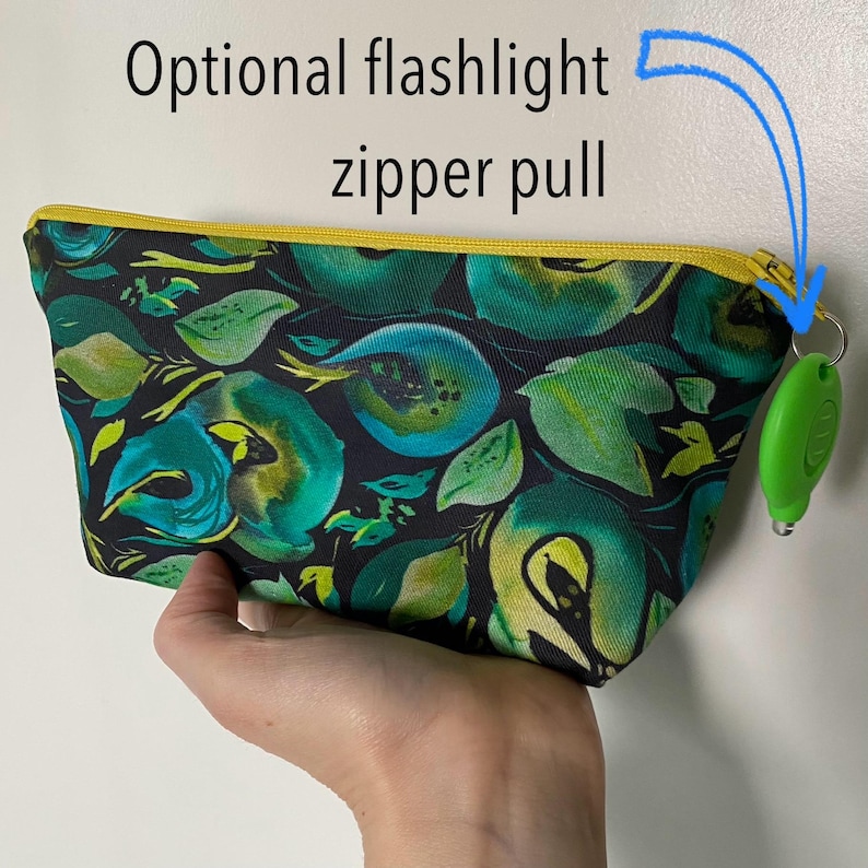 May include: A small, rectangular pouch with a floral pattern in shades of green and teal on a black background. The zipper is yellow, and the optional flashlight zipper pull is green. The text "Optional flashlight zipper pull" is above the pouch.