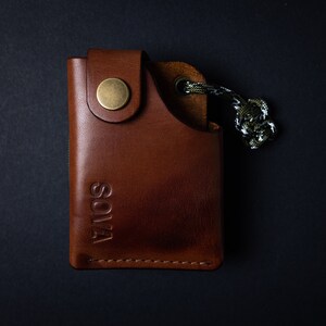 Peut inclure: Un portefeuille en cuir marron avec une fermeture &agrave; bouton en laiton et le mot "SOLVA" grav&eacute; sur le devant. Le portefeuille a une petite boucle avec un cordon vert et blanc attach&eacute;.