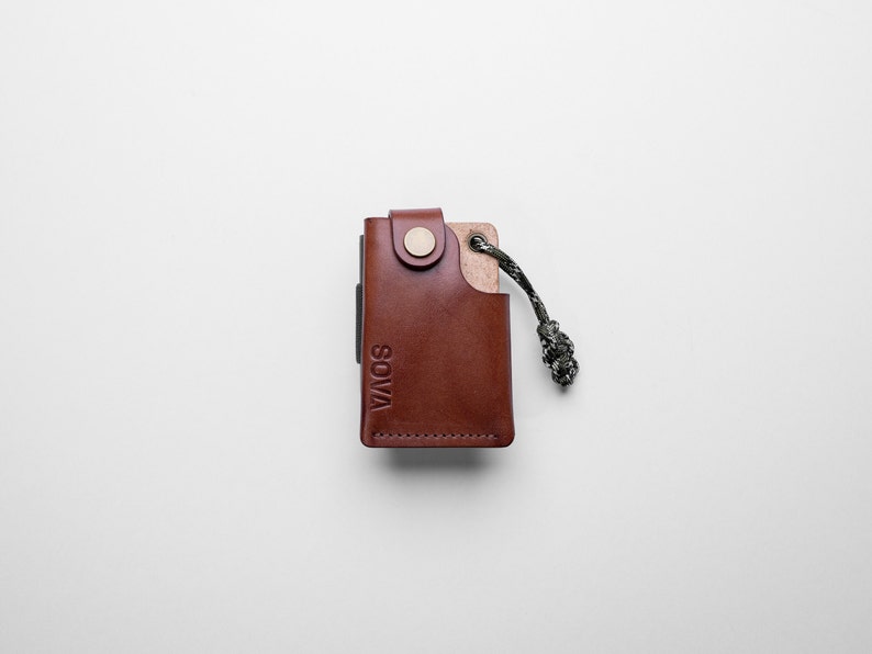 Peut inclure: Un portefeuille en cuir marron avec une fermeture &agrave; bouton m&eacute;tallique et le mot "SOVA" en relief sur le devant. Le portefeuille a une petite boucle en cuir marron avec un cordon noir et gris attach&eacute;.