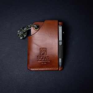 Peut inclure: Un portefeuille en cuir marron avec un cordon de paracorde vert et noir. Le portefeuille a une poche pour un stylo et un logo qui dit "SOVA HANDCRAFTED EST. 2018".