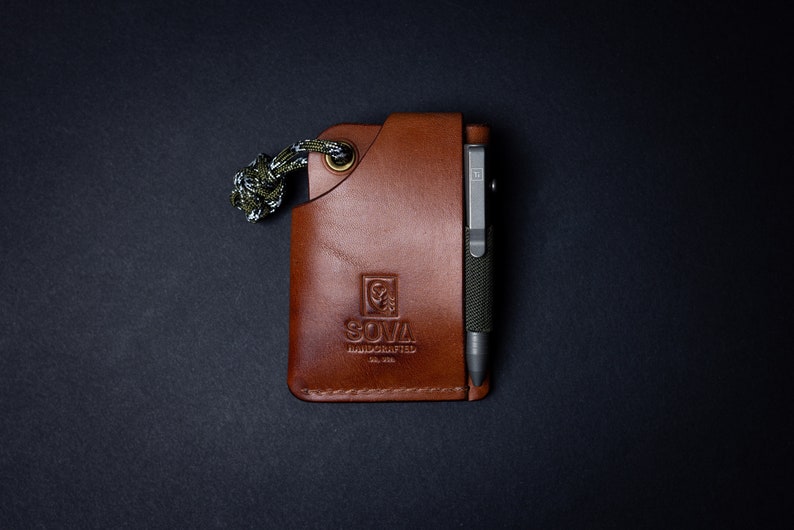 Peut inclure: Un portefeuille en cuir marron avec un cordon de paracorde vert et noir. Le portefeuille a une poche pour un stylo et un logo qui dit "SOVA HANDCRAFTED EST. 2018".