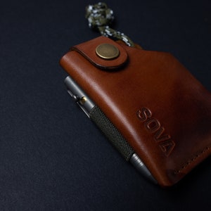 Peut inclure: Un portefeuille en cuir marron avec un stylo en m&eacute;tal et le mot "SOVA" en relief sur le devant.