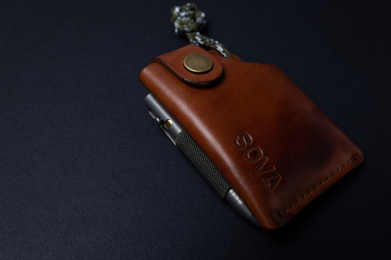 Peut inclure: Un portefeuille en cuir marron avec un stylo en m&eacute;tal et le mot "SOVA" en relief sur le devant.