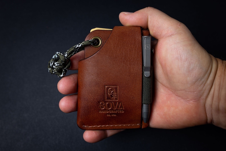 Peut inclure: Un portefeuille en cuir marron avec un porte-stylo et un stylo noir. Le portefeuille a un bord cousu et un logo qui dit "SOVA HANDCRAFTED" dans un carr&eacute;. Le portefeuille a un cordon vert et blanc attach&eacute; &agrave; lui.