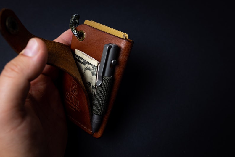 Peut inclure: Un portefeuille en cuir marron avec une sangle, un stylo et un billet de dollar &agrave; l'int&eacute;rieur. Le portefeuille porte un logo "SOMA".