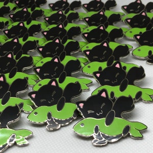 May include: A collection of black and green enamel pins featuring a black cat with pink ears and a white face, curled up on a green fish. The pins are silver-toned and have a shiny finish.